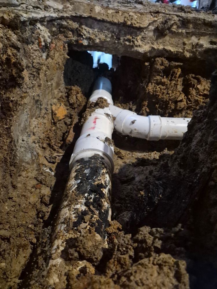 Broken underground PVC pipe in a dirt trench with white elbow fittings and muddy soil.