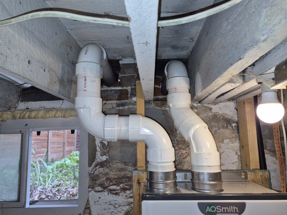 White PVC plumbing pipes and fittings under a floor, with a hanging bulb and window opening nearby.