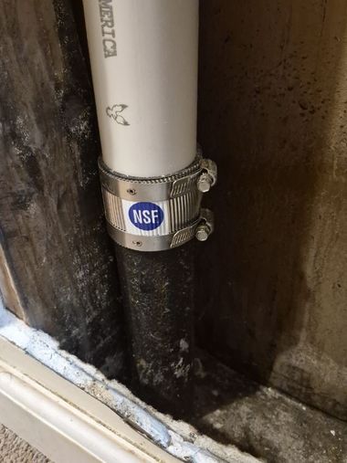White PVC pipe with NSF clamp in a damp, moldy corner beside a wall and baseboard