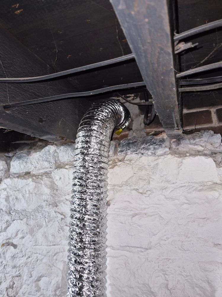 Flexible silver duct venting through a hole in a dark floor or ceiling, against a white wall