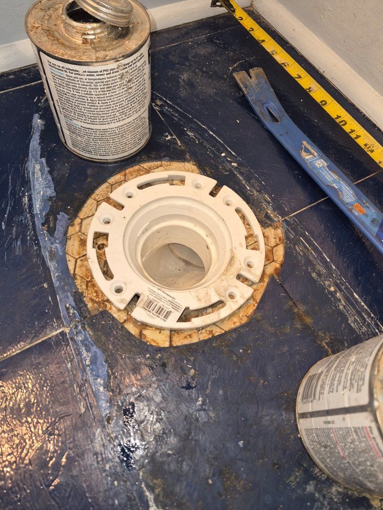 Plumbing parts on a dark floor, including a white toilet flange, metal canister, and blue tool.