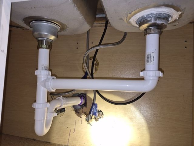 Under-sink plumbing with two drain pipes, a horizontal connector, and exposed wires in a wooden cabinet.
