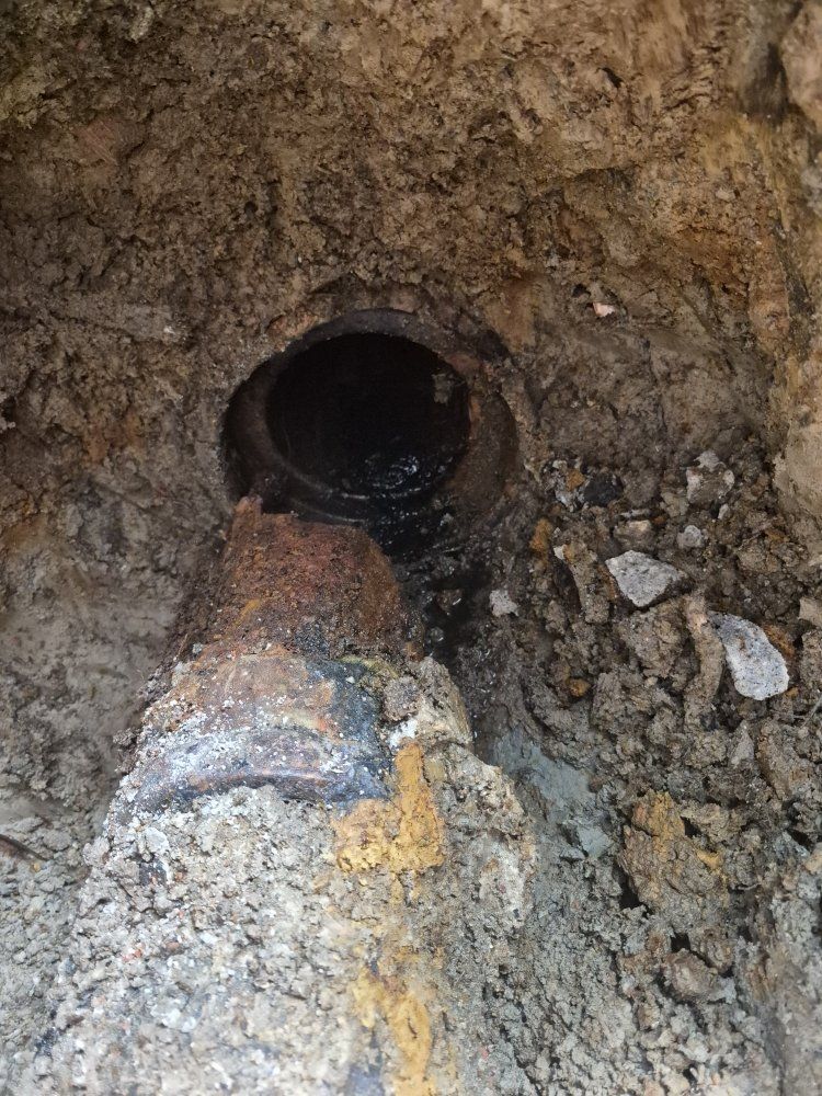 A rusty pipe opening in a muddy dirt wall, with rocks and gravel at the base