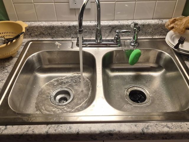 Double kitchen sink with running faucet, left basin draining, green sponge hanging on the right faucet.