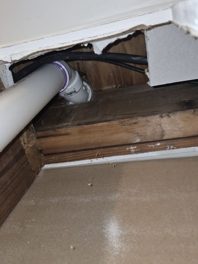 Exposed plumbing and a large pipe entering a damaged wall cavity above a floorboard opening