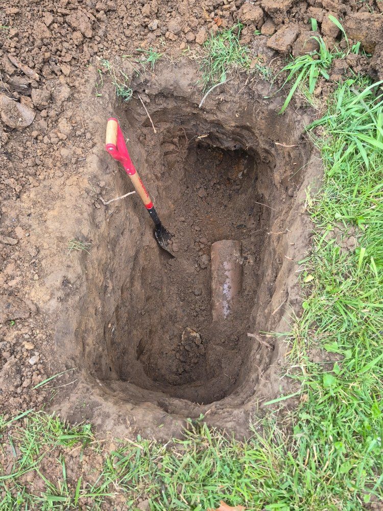 A red-handled shovel in a narrow dirt hole beside green grass and loose soil.