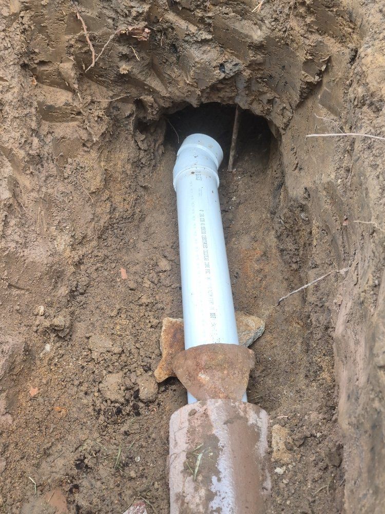 White PVC pipe in a dirt trench, connected to a rusted pipe fitting underground.