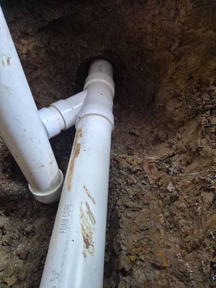 White PVC pipes in a dirt trench, showing a plumbing connection underground.