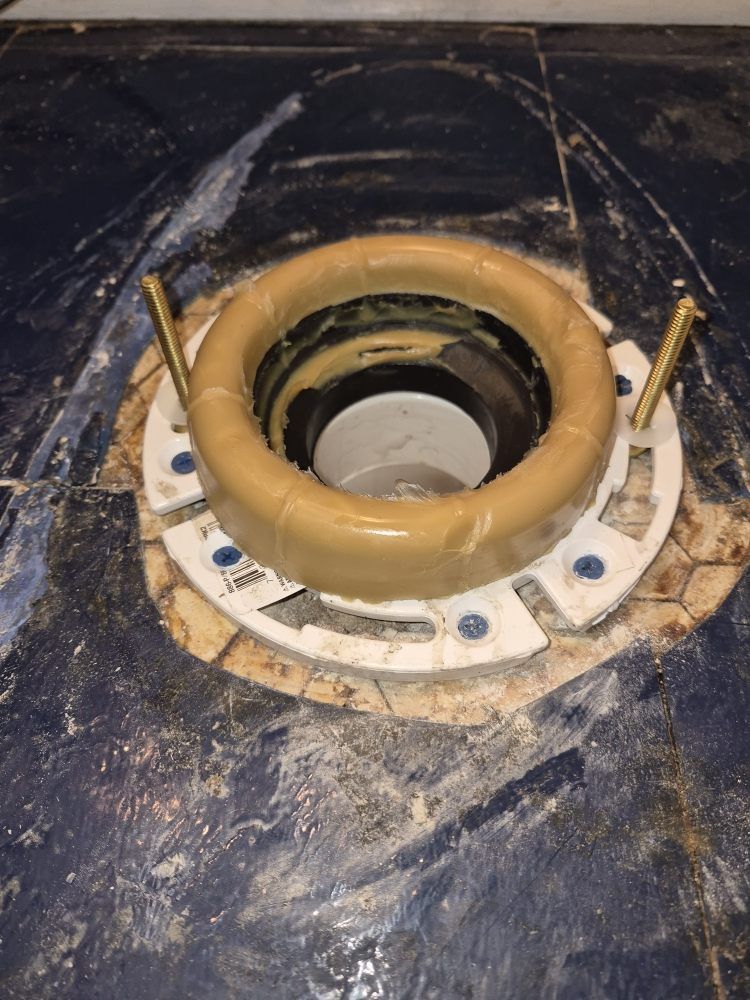 Close-up of a floor-mounted circular plumbing flange with brass bolts and a tan wax ring around a drain opening