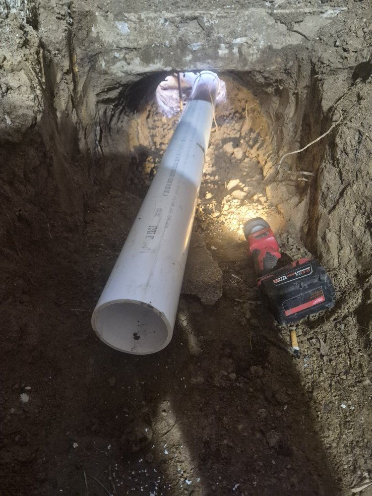 Worker installing a large gray pipe in a dug trench beside exposed soil and rock.