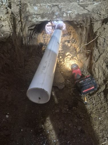 Long PVC pipe extending through a dirt trench into a hole in a concrete wall, with a cordless drill nearby