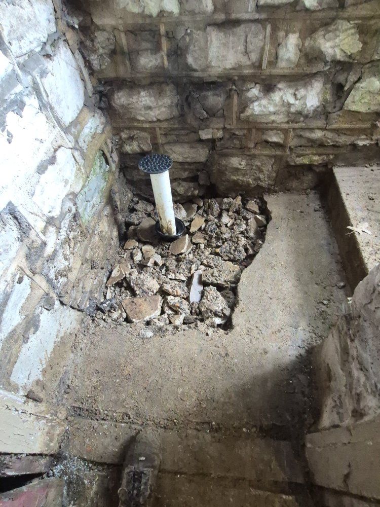 Rough basement corner with exposed stone walls, a floor drain pipe, and broken concrete around it