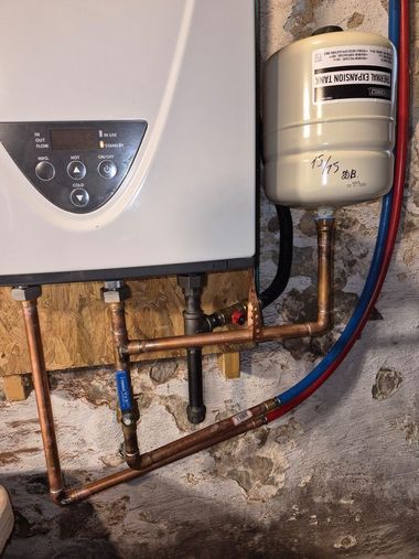 Wall-mounted boiler beside a white expansion tank with exposed copper pipes in a damp basement corner