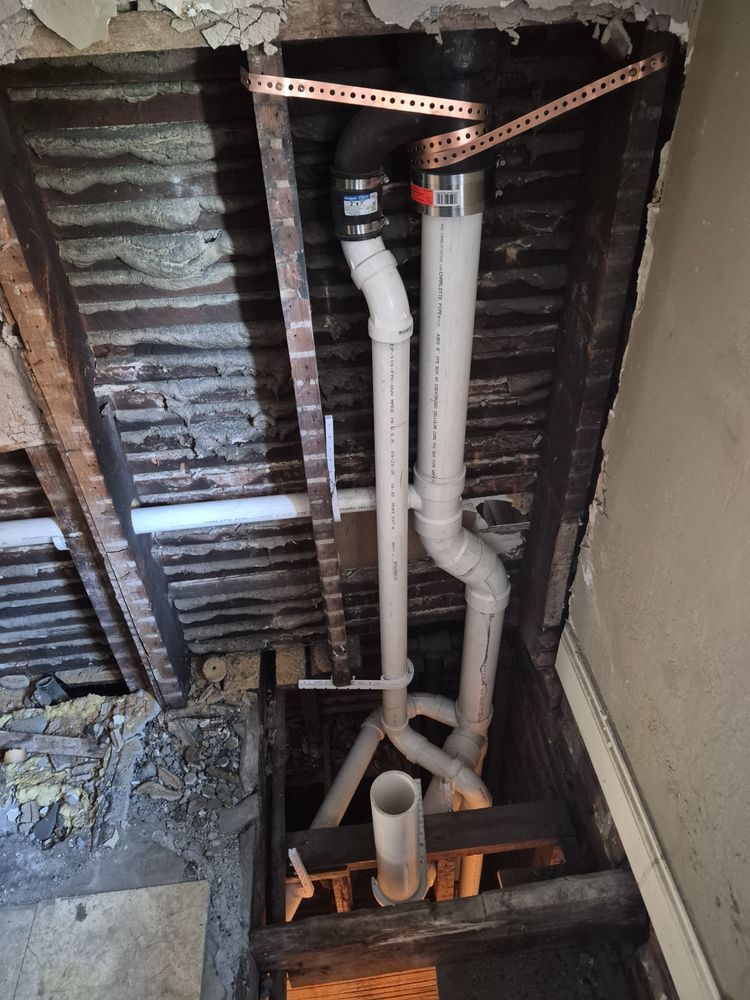 Exposed plumbing pipes in an unfinished ceiling, with insulation and a wall opening below.