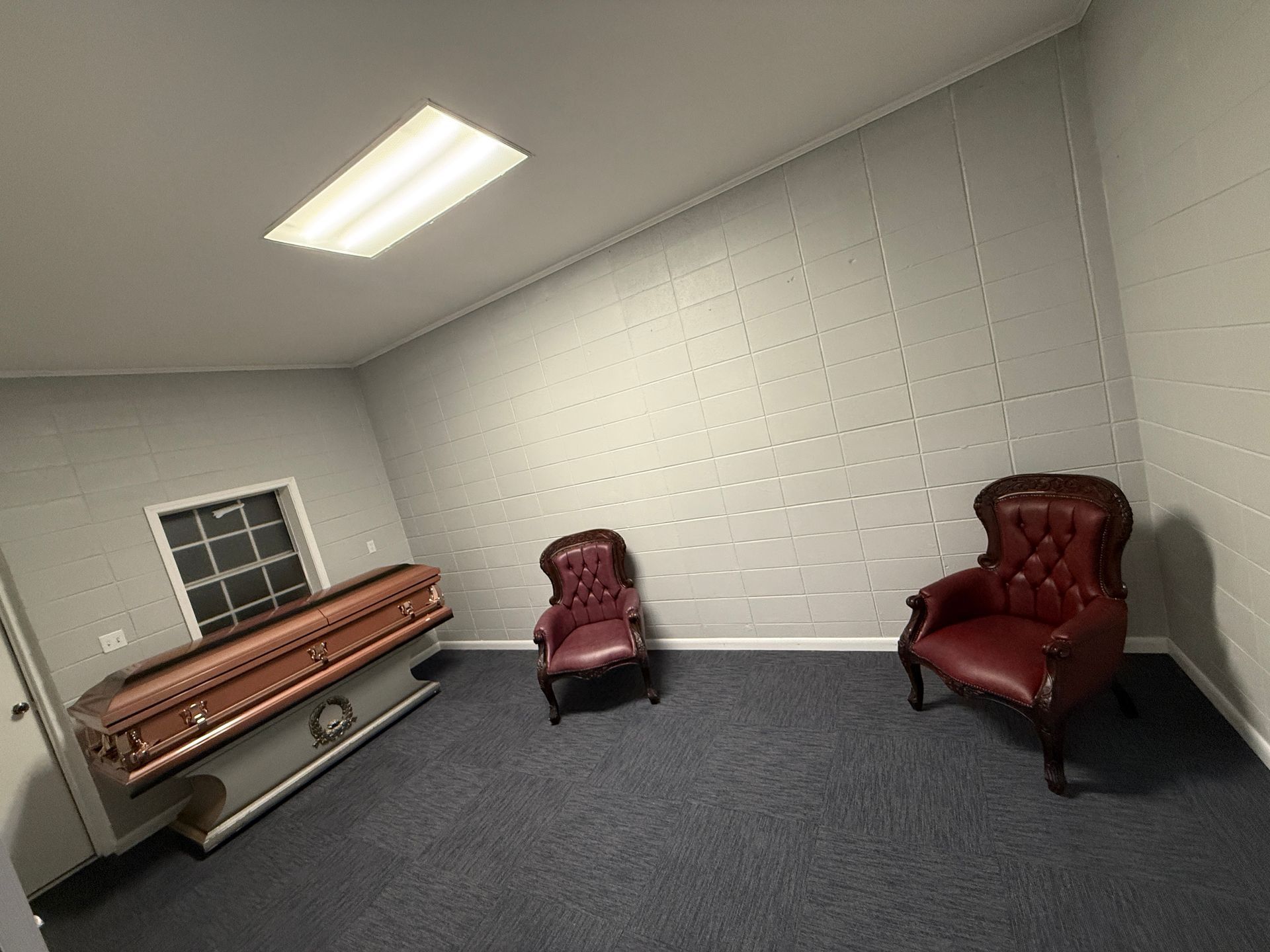 Small room with gray carpet, a casket near a window, and two red armchairs against the wall.