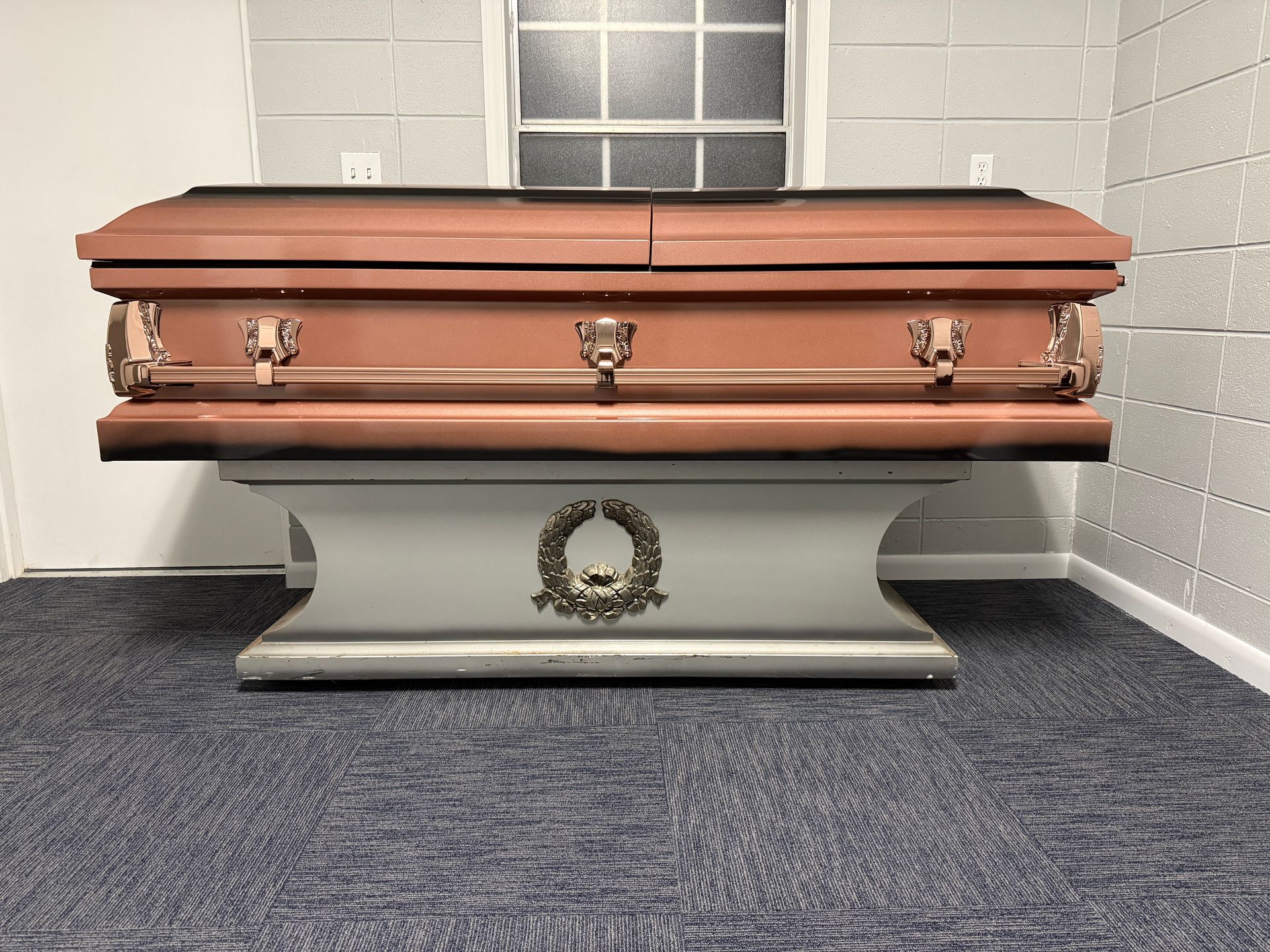 Copper casket on a stand in a tiled room with a window and wreath on the base