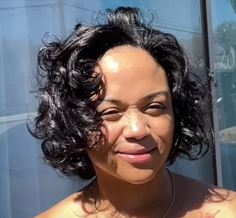 A woman with dark curly hair smiles at the camera in sunlight