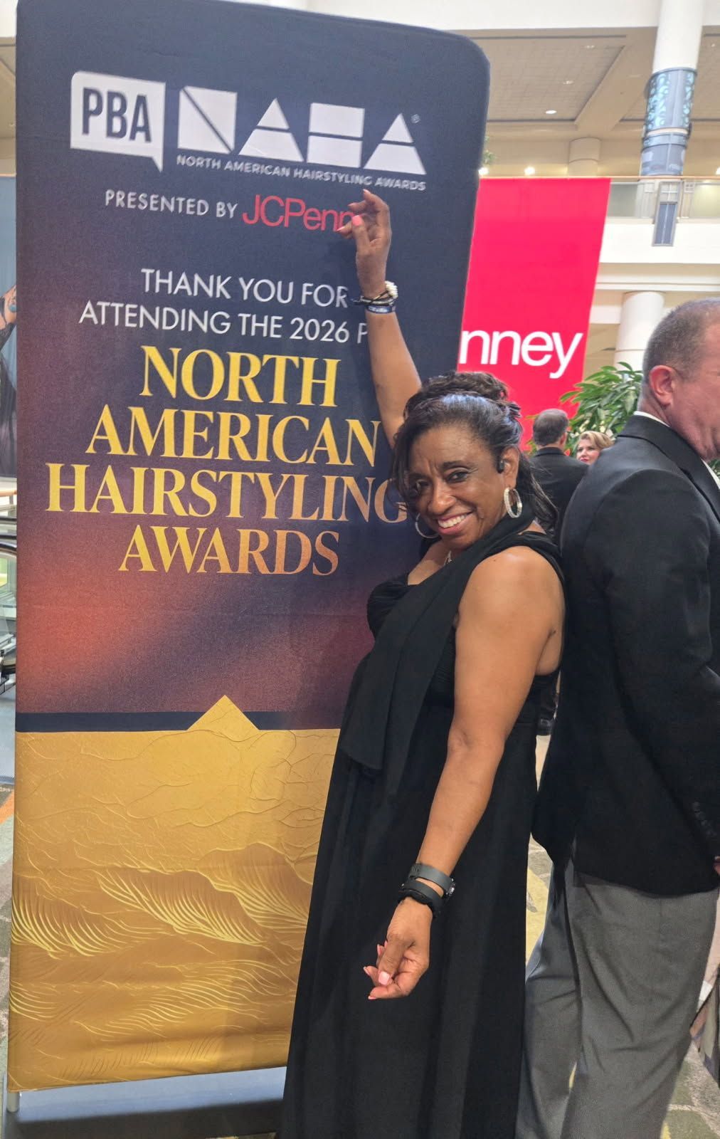 Woman in a black dress posing by a North American Hairstyling Awards banner, smiling at an event.