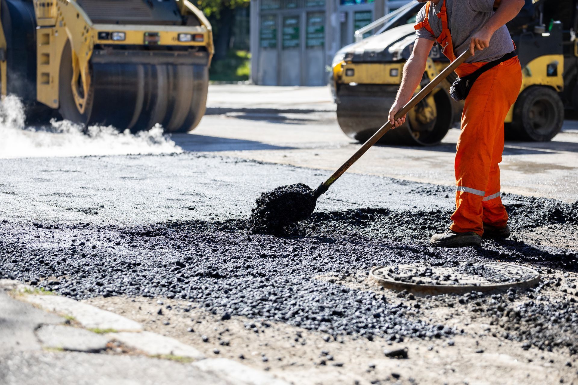 Worker in orange gear shovels fresh hot-mix onto a road beside a steamroller during asphalt repair.
