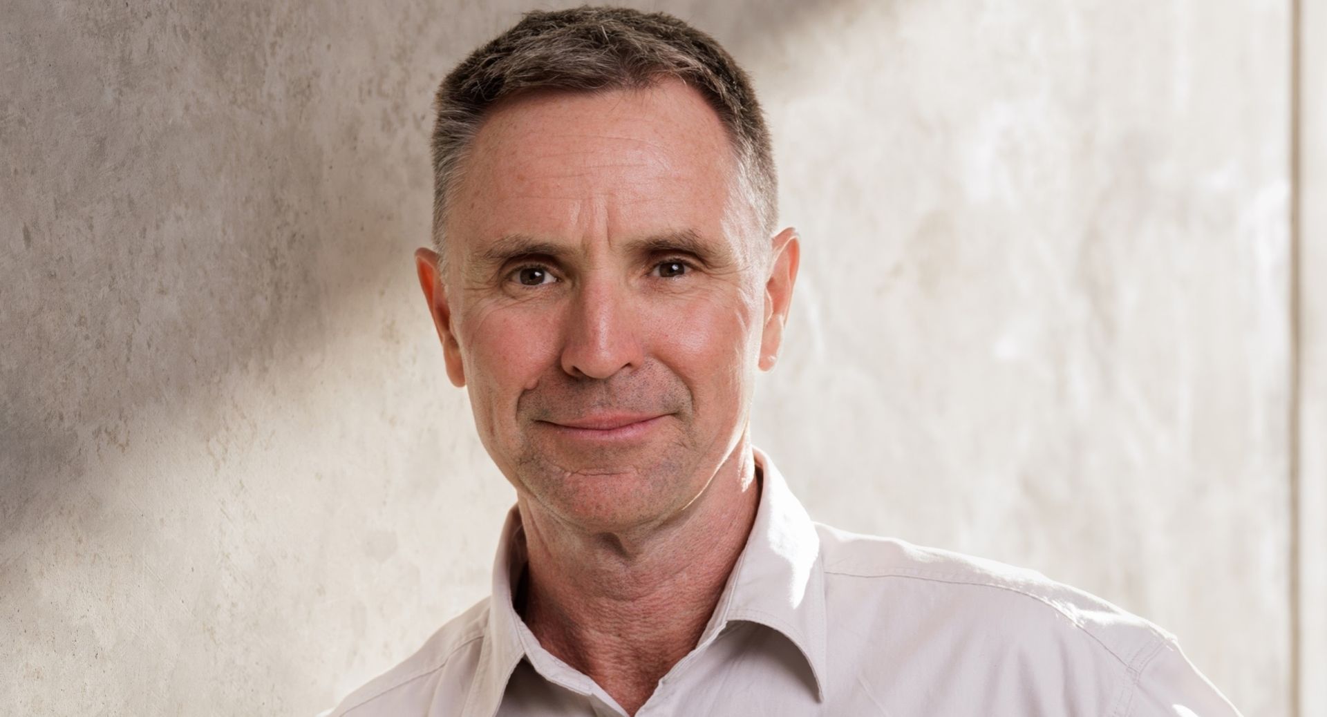 Portrait of a man in a white shirt against a light textured wall, looking at the camera.