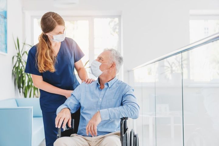 Young woman nurse explaining information to man patient in wheelchair in medical face mask while talking together in hospital.