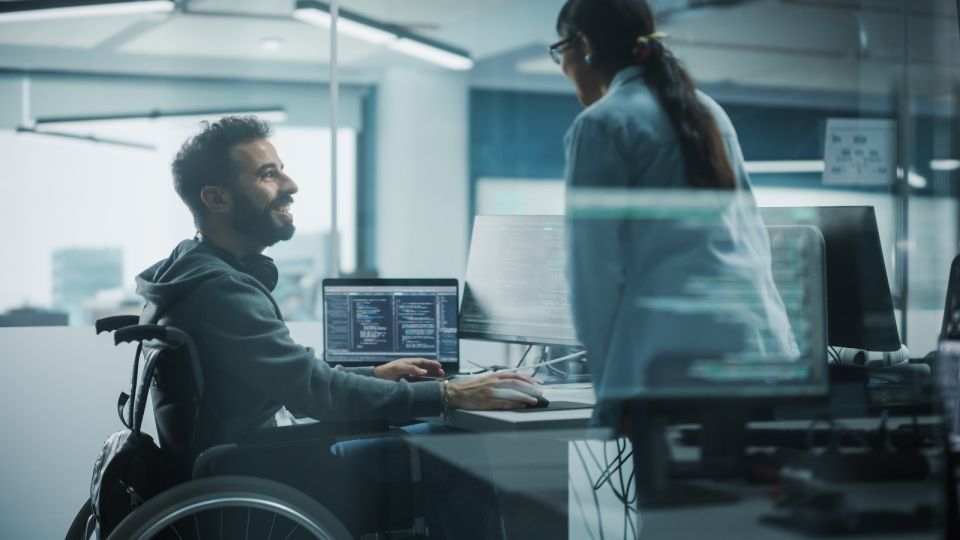 Disability in a Wheelchair Talks with Colleague while Working on Computer.