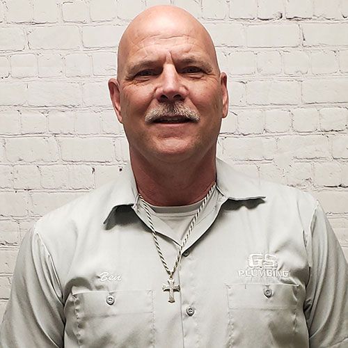 A person in a grey GS Plumbing work shirt and a cross necklace posing against a white brick wall.