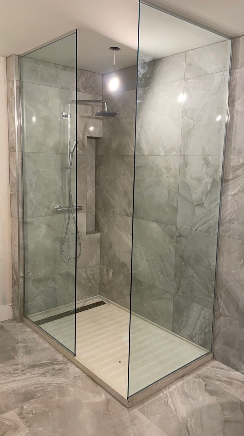 Une douche moderne aux parois de verre avec carrelage gris, pomme de douche et drain, à l'intérieur.