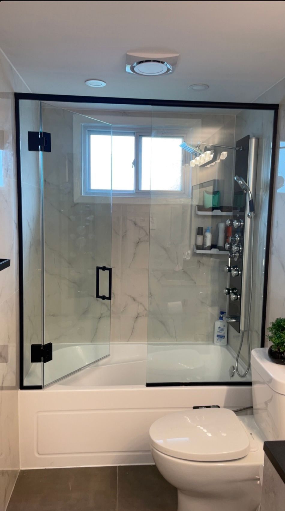 Salle de bain avec porte de douche en verre, garniture noire, baignoire blanche, toilettes et fenêtre.