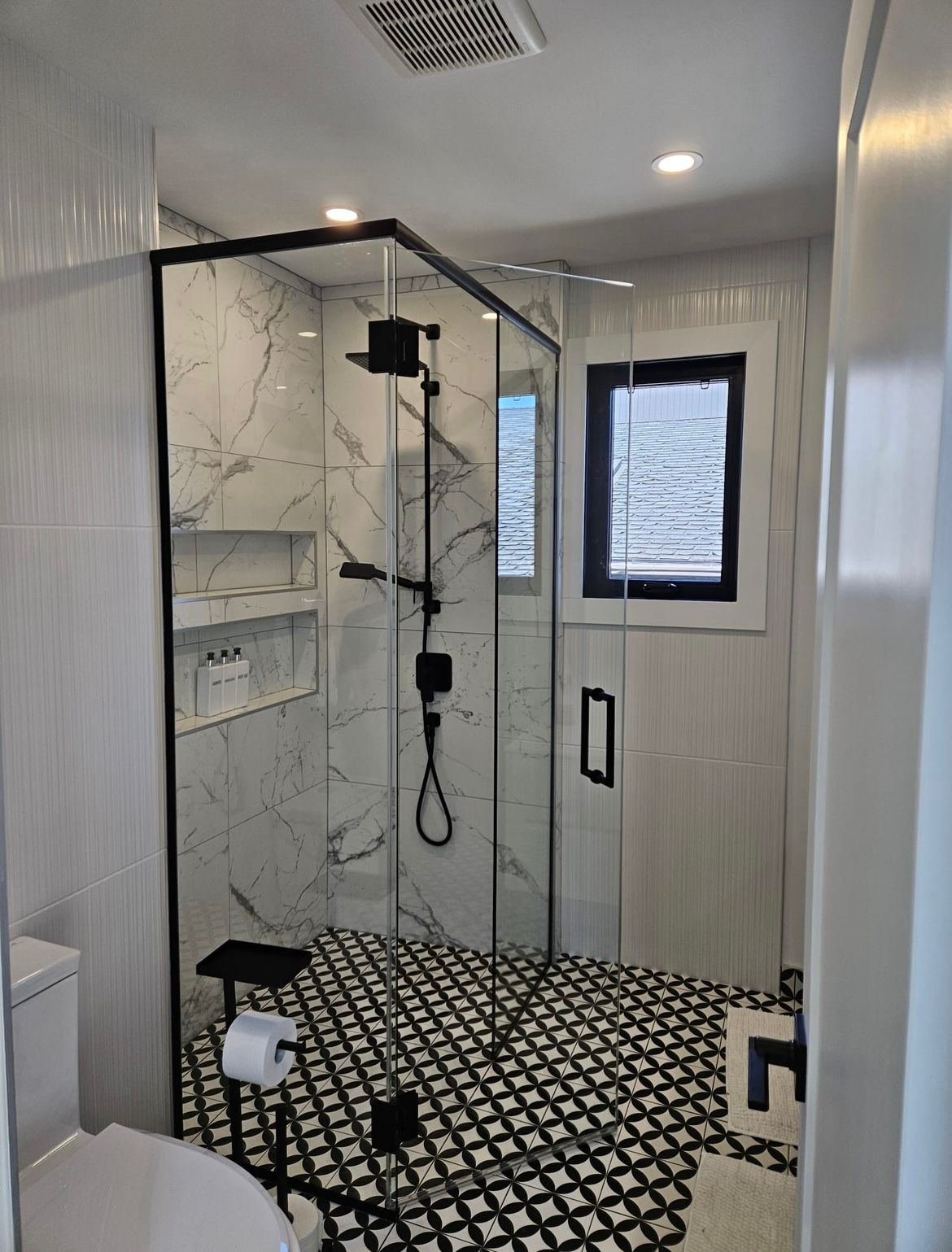 Salle de bain moderne : douche carrelée en marbre avec des touches noires, sol à motifs noirs et blancs, toilettes et petite fenêtre.