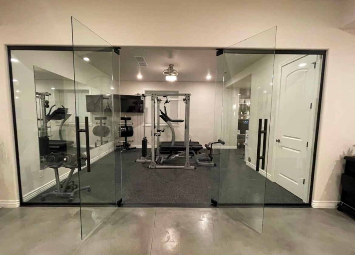 Salle de sport à domicile aux parois vitrées, avec équipements de sport à l'intérieur. Sol gris, cadre noir, murs blancs et porte blanche.