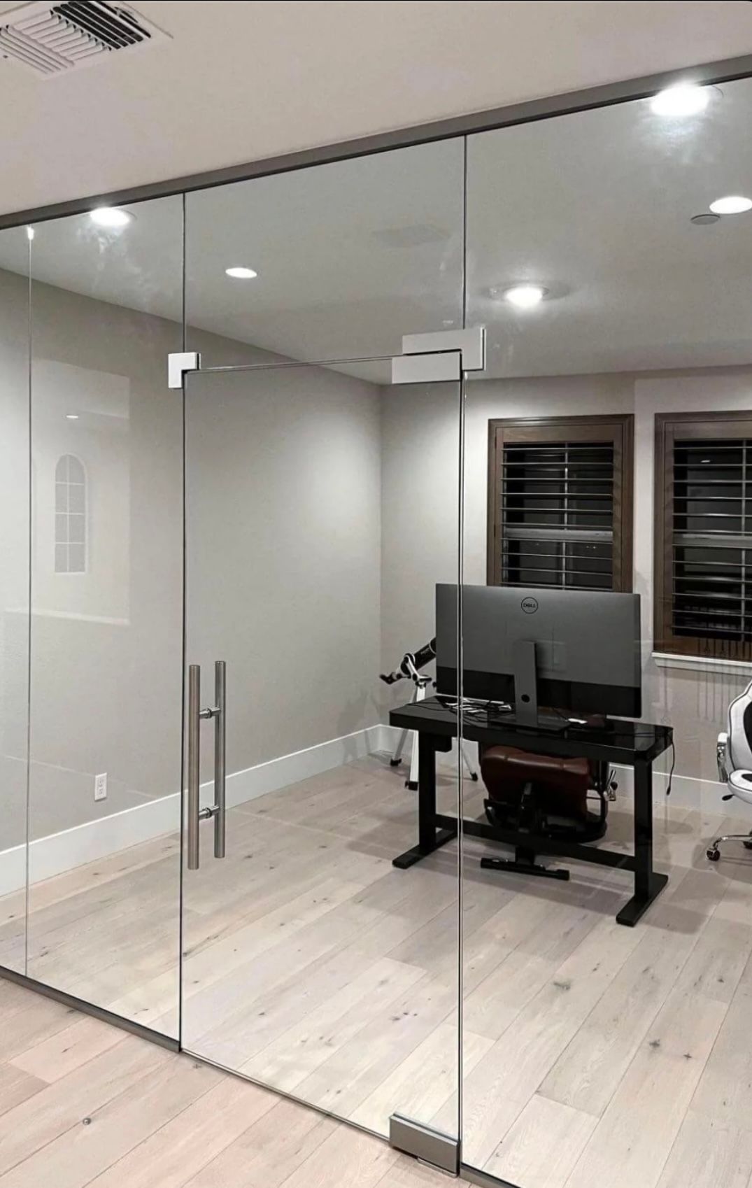Bureau à domicile aux parois vitrées avec ordinateur, bureau, stores en bois et porte avec poignée en acier inoxydable.