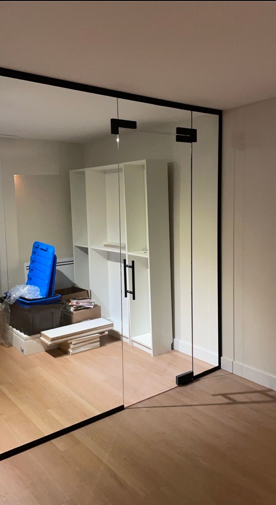 Une porte et un mur en verre à cadre noir ferment une pièce au parquet. Des étagères blanches et un peu de désordre sont visibles à l'intérieur.