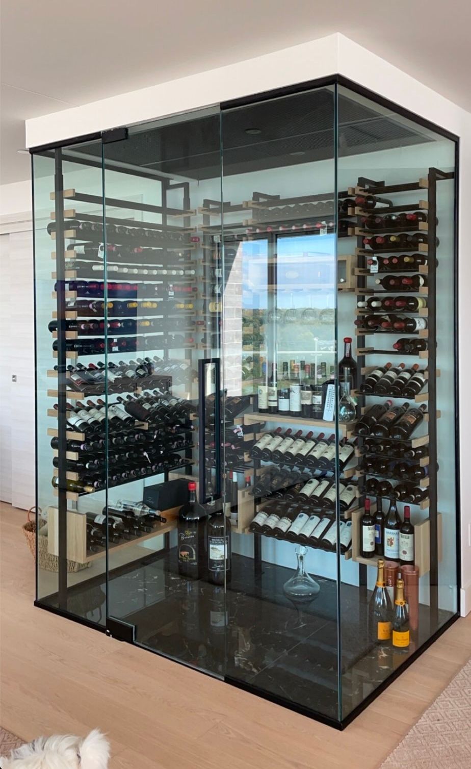 Cave à vin vitrée avec rangées de bouteilles exposées. Étagères en bois et métal contre les parois vitrées.