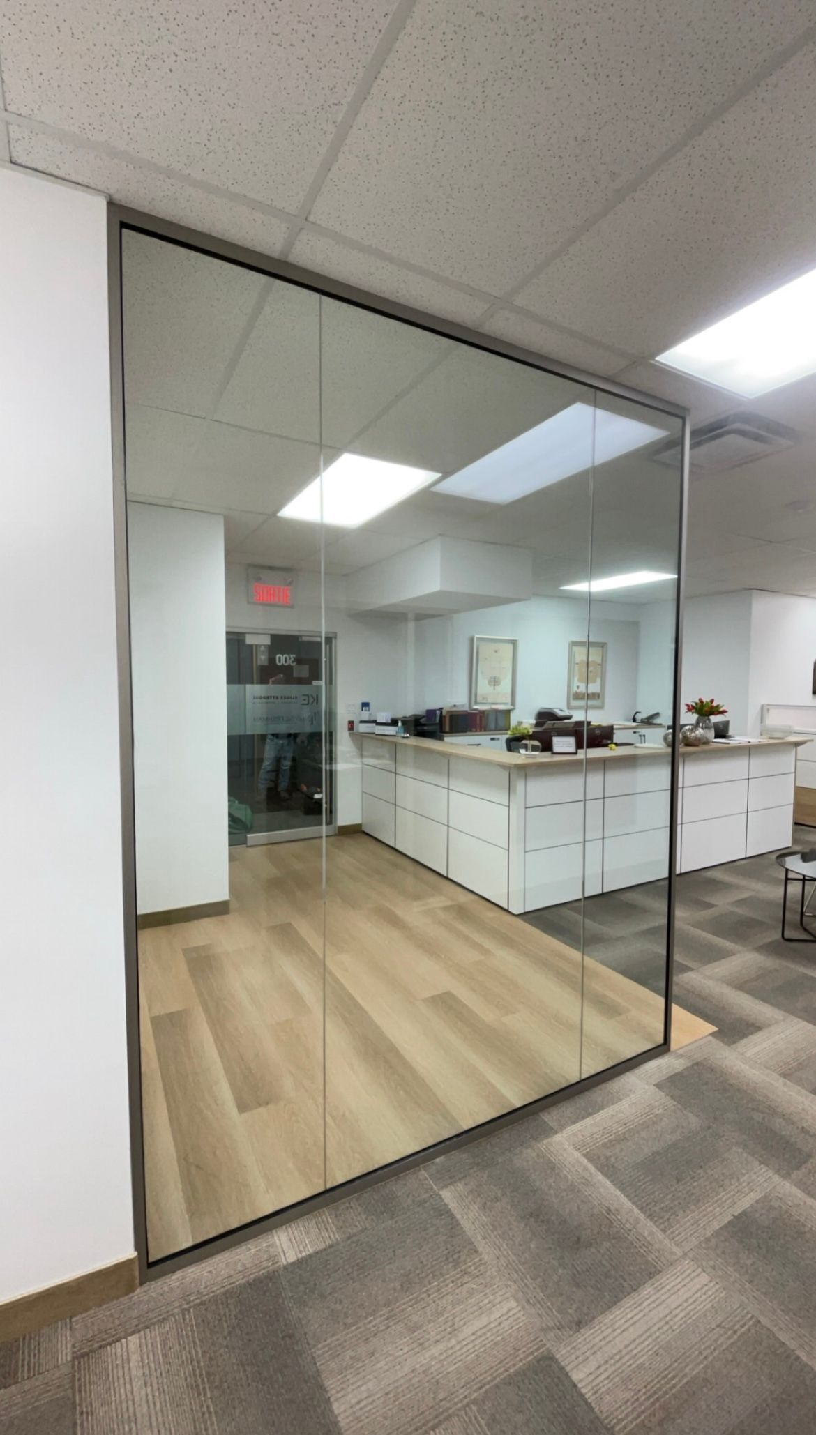 Cloison de bureau en verre avec cadre en acier inoxydable, reflétant l'espace de travail intérieur avec des armoires blanches et des planchers en bois beige.