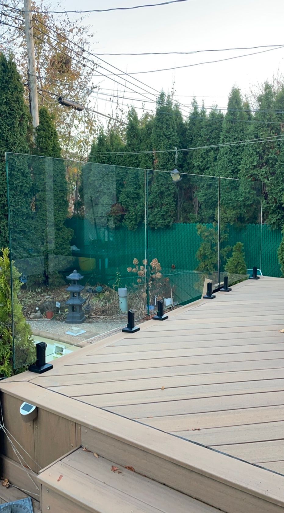 Terrasse avec garde-corps en verre donnant sur une cour arrière avec clôture verte et arbres.