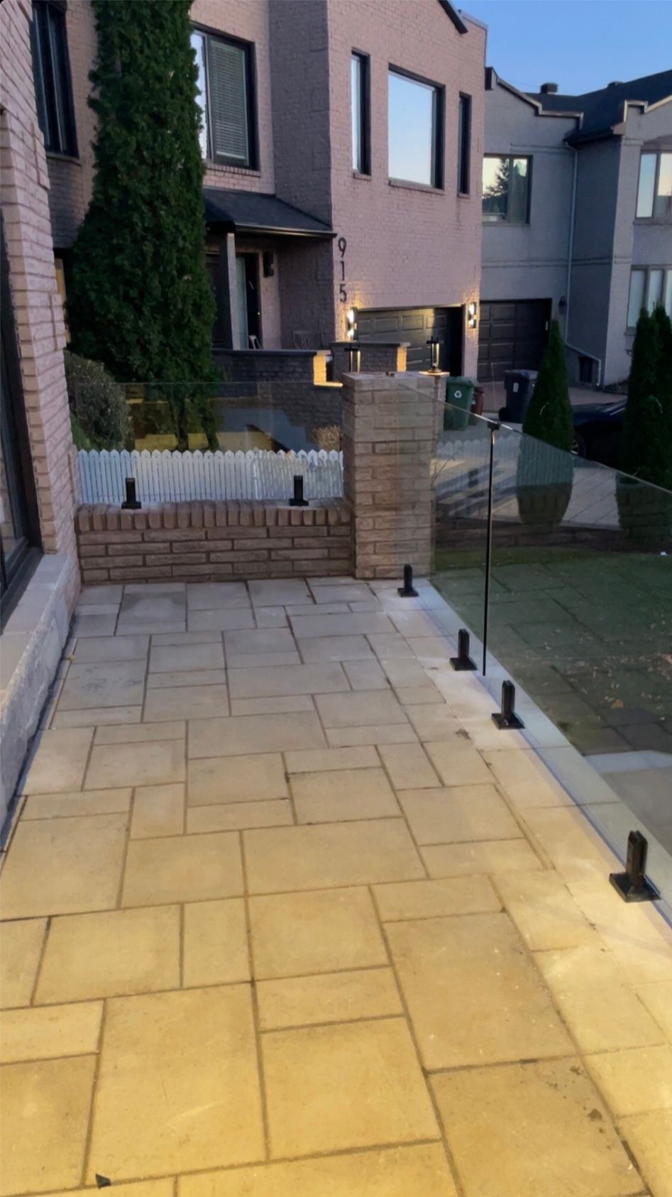 Patio en pierre avec balustrade en verre donnant sur une cour herbeuse; mur de briques adjacent, bâtiments beiges en arrière-plan.