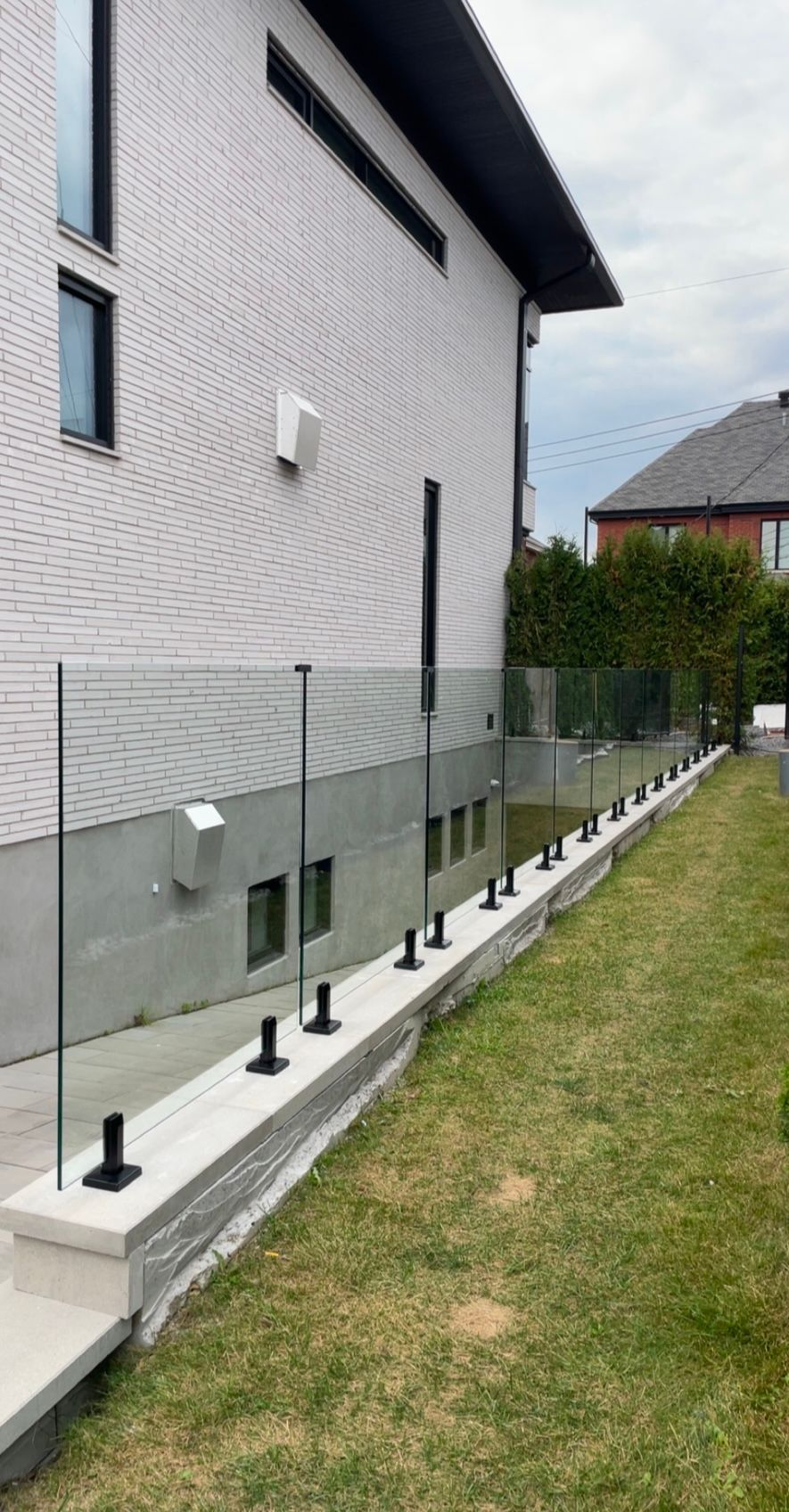 Clôture en verre le long d'un mur de soutènement en béton à côté d'une zone herbeuse, avec un bâtiment en briques blanches derrière.