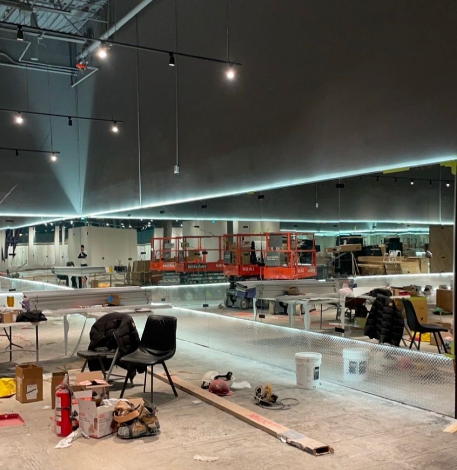 Chantier en cours, grande pièce vide, plafond inachevé avec lumières, matériel de travail, ascenseur orange, chaises noires.