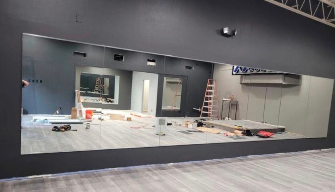 Un grand miroir sur un mur noir reflète une pièce inachevée, des outils et une échelle dans une salle de sport.