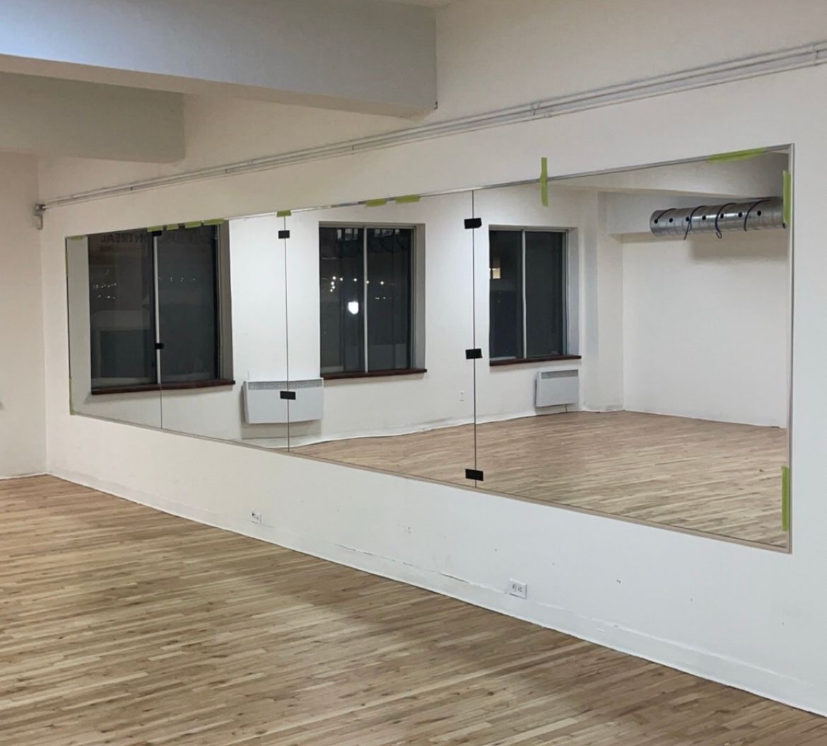 Studio de danse avec de grands miroirs sur un mur, des fenêtres et un plancher en bois.
