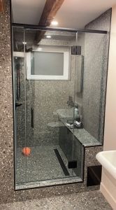 Douche en verre avec garniture noire, carrelage gris moucheté, un lavabo et une petite fenêtre.