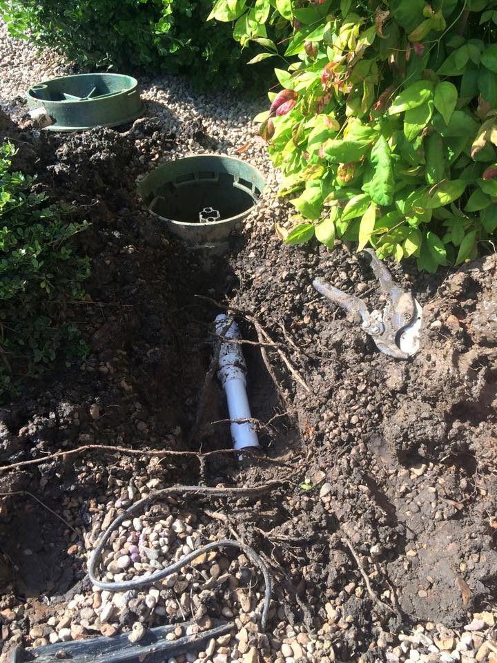 Garden trench with exposed irrigation pipe and two green utility covers beside shrubs
