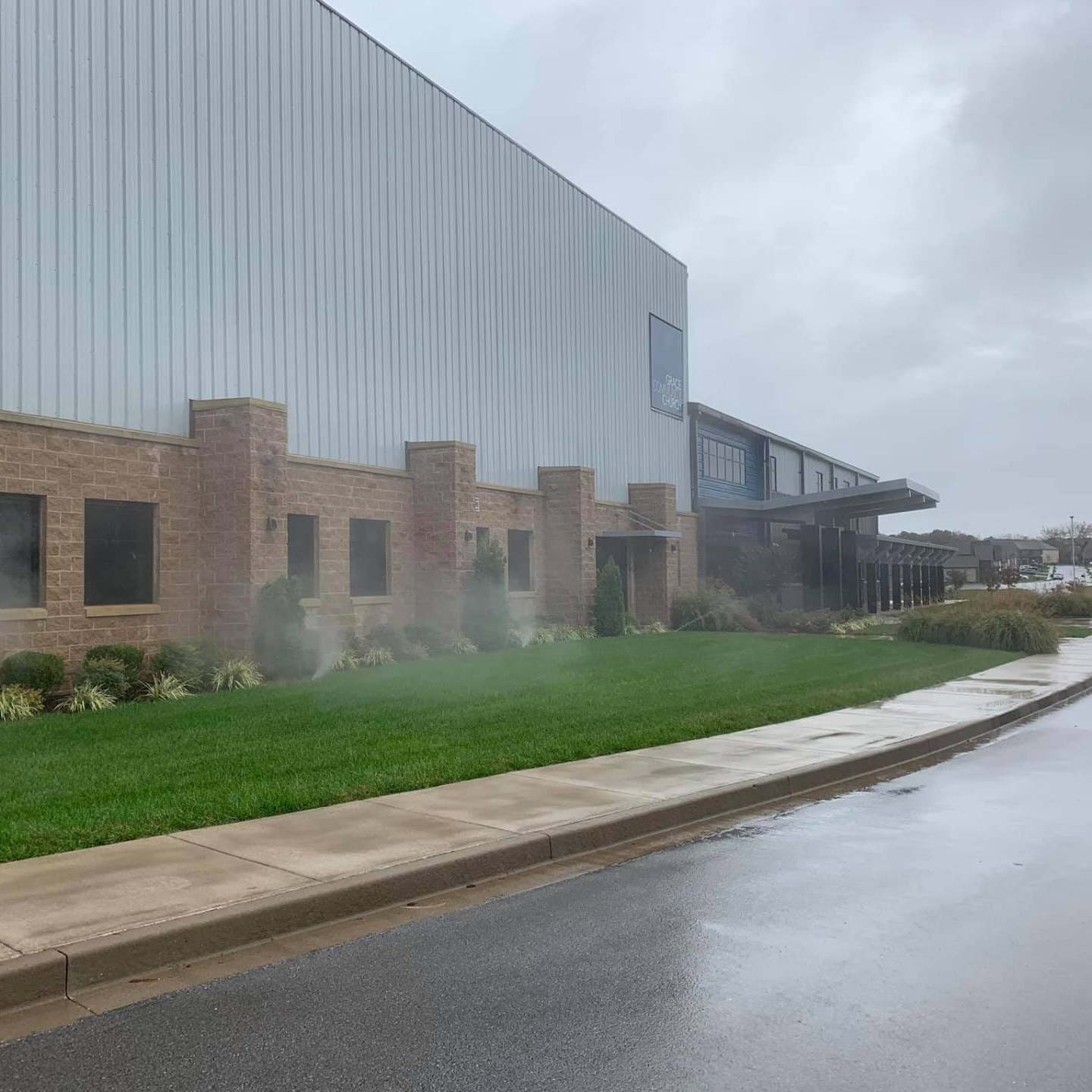 Modern building with brick and metal siding beside a wet road and green lawn on a cloudy day