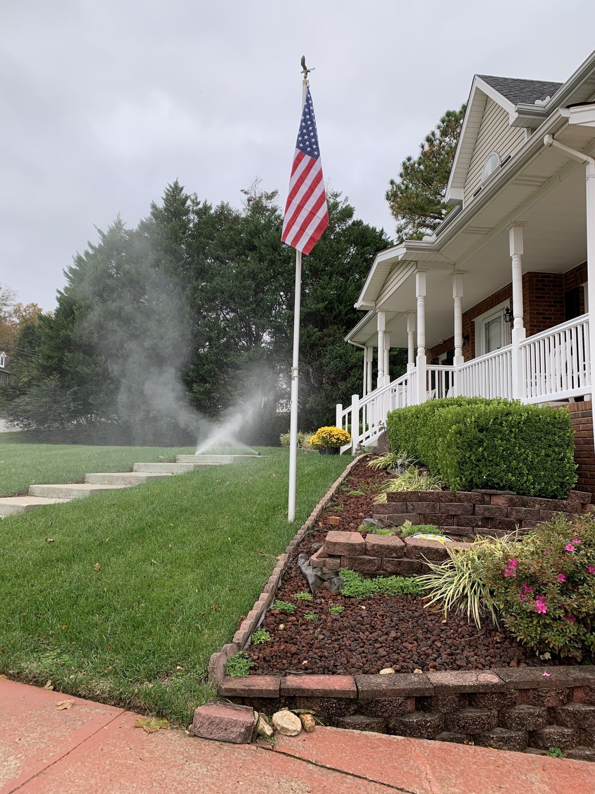 American flag by a white house with porch, front garden, and sprinklers watering the lawn