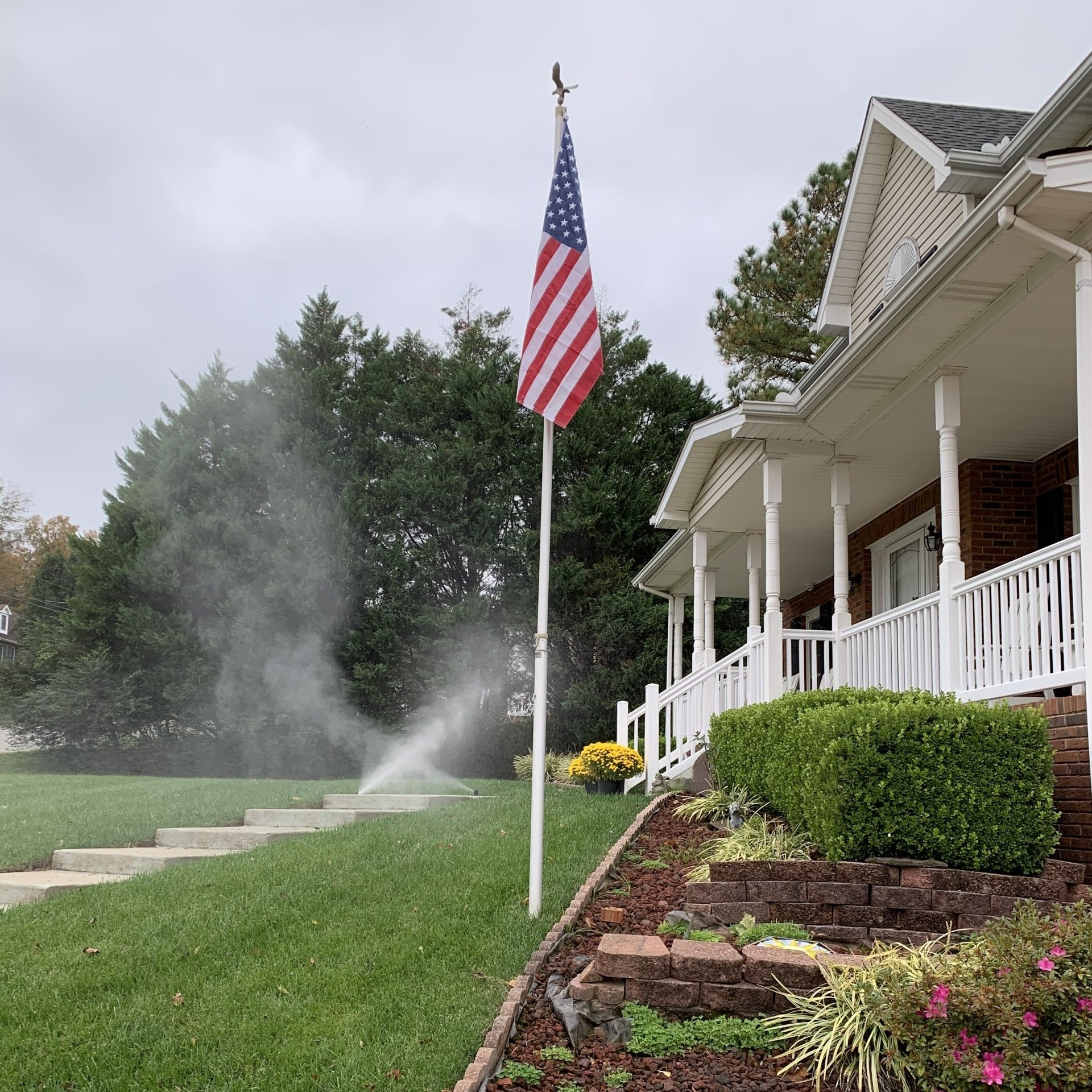 American flag by a suburban house with porch, lawn sprinklers spraying, and flower beds in front