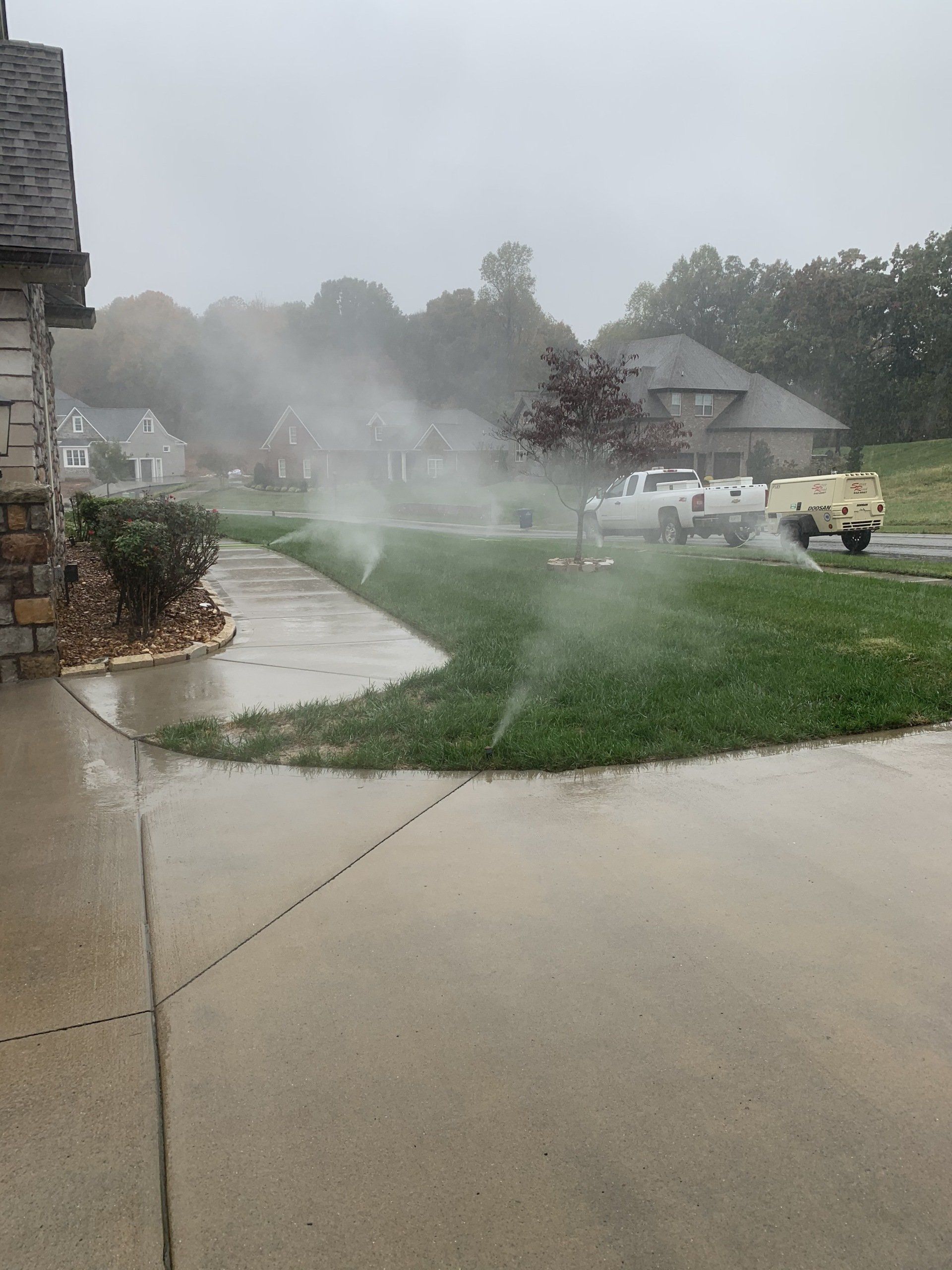 Rainstorm spraying a suburban street and lawn, with mist over sidewalks and parked vehicles.