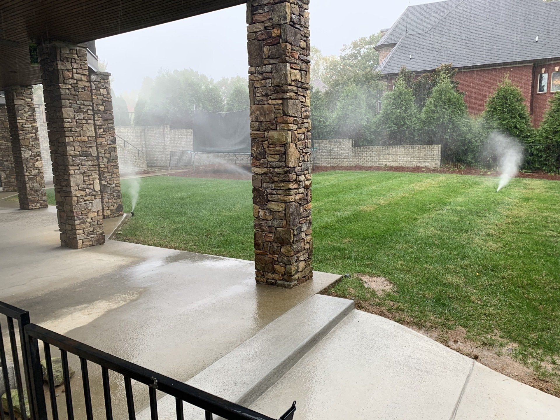 Covered patio overlooking a misty backyard with green lawn, stone columns, and sprinkler spray.