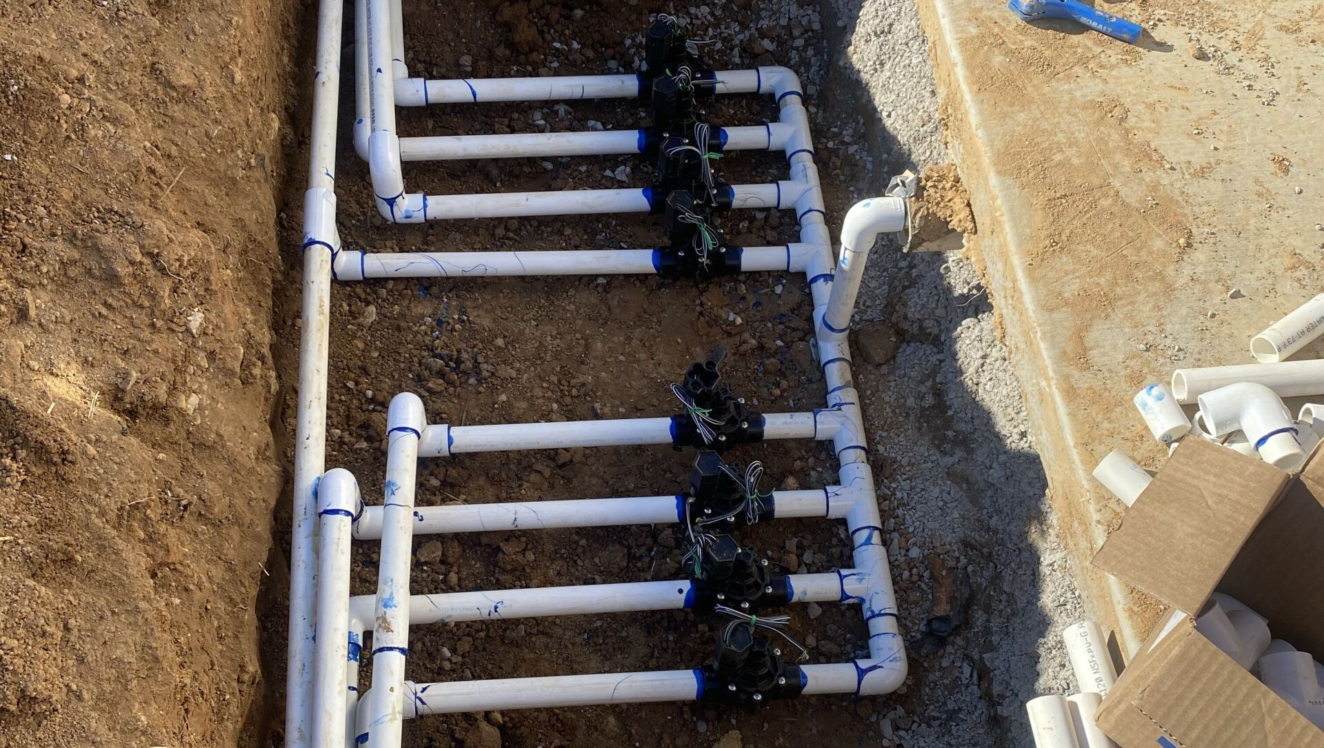 Open trench with white PVC pipes and black fittings laid in a row for plumbing installation