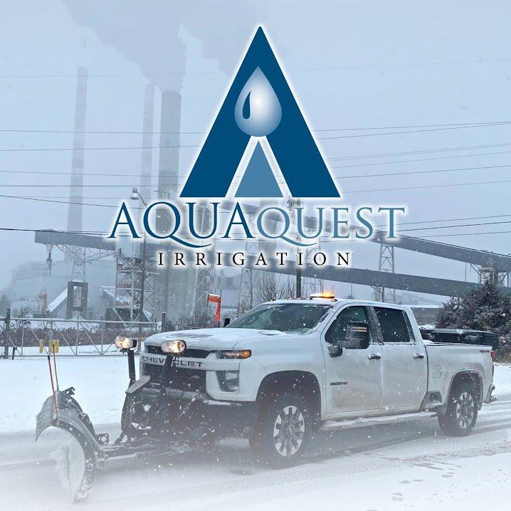 White pickup truck with snowplow in snowy field beside AquaQuest Irrigation sign and bridge overhead